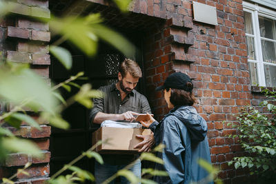 Mature man doing electronic signature while receiving parcel from female delivery person standing at doorstep