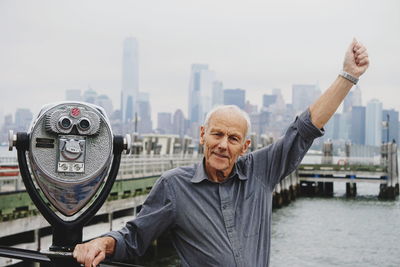 Portrait of senior man standing by coin-operated binoculars against river in city