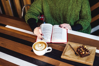 Midsection of woman with coffee and cake on table reading book while sitting in cafe