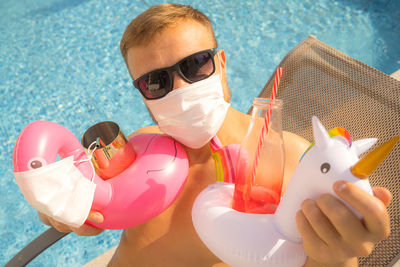 High angle portrait of man wearing mask holding drinks and inflatable rings on pool
