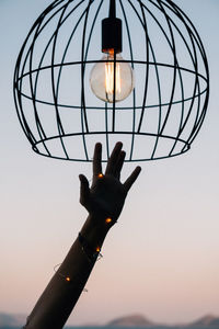 Low angle view of hand by illuminated light bulb against sky during sunset
