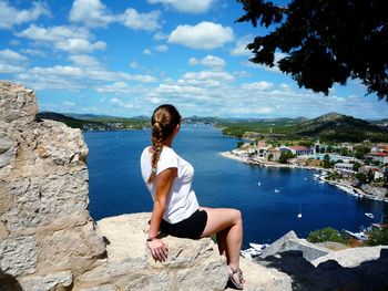 Woman looking at view while sitting on rock by sea against sky