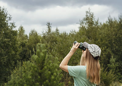 Young blonde woman bird watcher in cap  looking through binoculars sky in summer forest ornithology