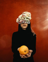 Woman with eyes covered by flowers holding fruit against wall