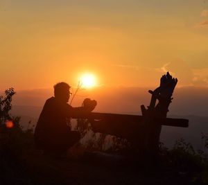 Silhouette man crouching while looking at mountains against orange sky during sunset