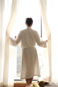 Rear view of woman in bathrobe looking out through window at home