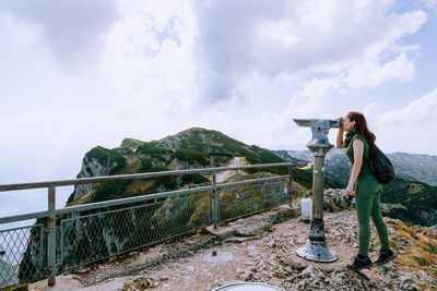 Side view of woman looking through telescope while standing on mountain against sky