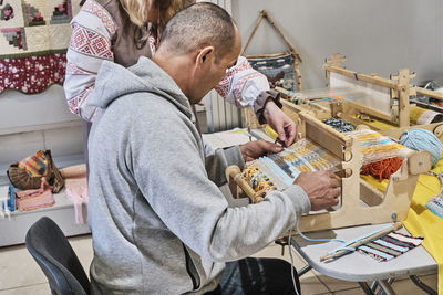 Faceless artisan woman teaching senior asian man to weaving small rug on table loom.