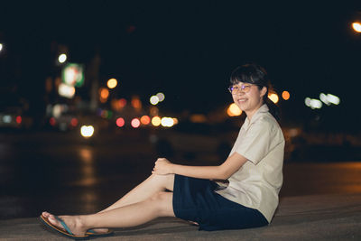 Portrait of smiling young woman sitting in city at night