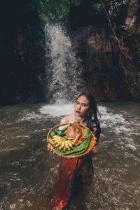 Portrait of young woman holding plate while standing in river against waterfall