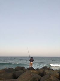 Man fishing on rock by sea against sky