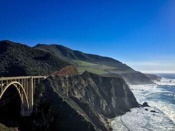 Bixby creek bridge at rocky mountains by sea against blue sky
