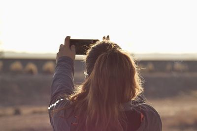 Rear view of woman photographing with smart phone against clear sky