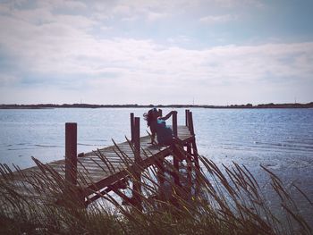 Woman sitting on pier over lake against cloudy sky