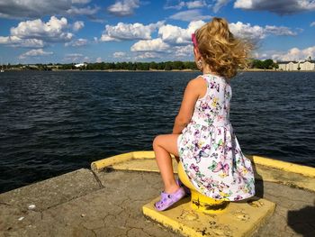 Girl sitting on cleat at river against sky