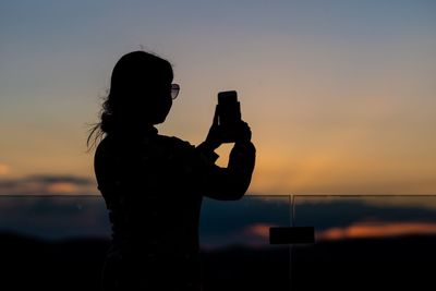 Silhouette woman photographing orange camera against sky during sunset