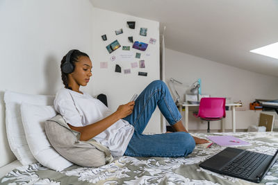 Young girl listening music while using smart phone sitting at home in bedroom