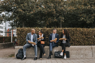 Male and female corporate colleagues having lunch while sitting on bench near plants at sidewalk