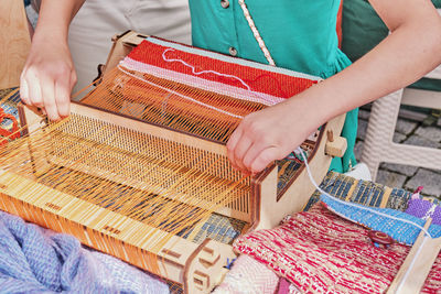 Hands of  girl weaving small rug with pattern on manual table loom at outdoors workshop on weaving. 