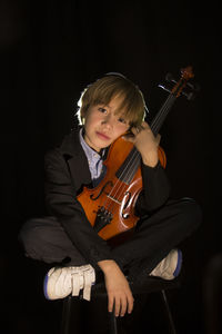 Boy holding violin while sitting against black background