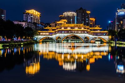 Illuminated bridge over river by buildings against sky at night