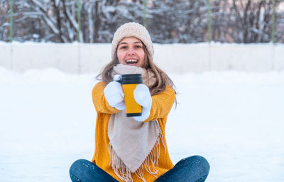 Portrait of happy young woman with ice skates sitting on the snow and holding mug with coffee