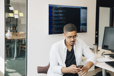 Male computer hacker using smart phone while sitting at desk in creative office