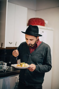 Businessman wearing hat having food while standing by kitchen counter at creative office