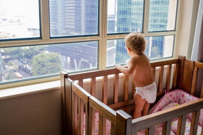 Rear view of shirtless boy looking through window at home