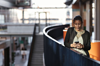 Mid adult businesswoman using mobile phone while standing at railing in office lobby