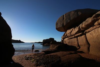 Rear view of woman by rock formation at beach against clear blue sky