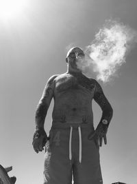 Low angle view of shirtless man smoking while standing against sky