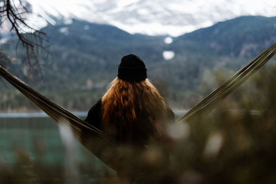 Back view of serene female traveler sitting in hammock and enjoying majestic view of lake in mountains in autumn