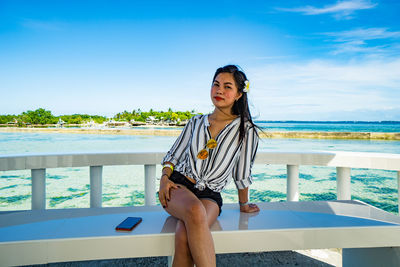 Young woman sitting by sea against blue sky