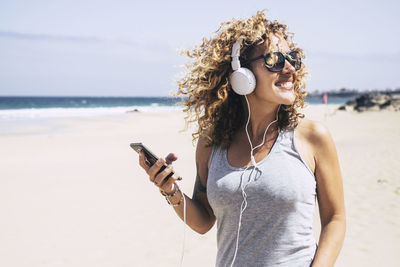 Smiling woman listening music while standing at beach during summer