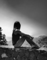 Young woman sitting on retaining wall against sky