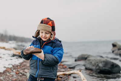 Smiling boy looking at rock while standing on shore against clear sky