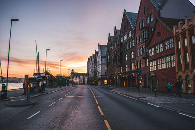 Empty road by buildings against sky during sunset