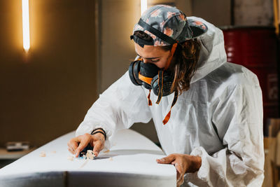 Side view of male shaper in safety costume and respirator polishing surface of surf board with hand plane in workshop