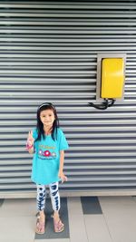 Portrait of smiling girl showing peace sign against corrugated wall