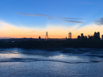 Silhouette buildings by sea against sky during sunset