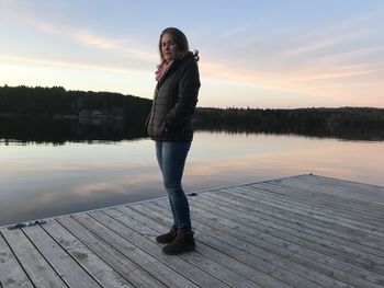 Woman standing on pier at lake against sky during sunset