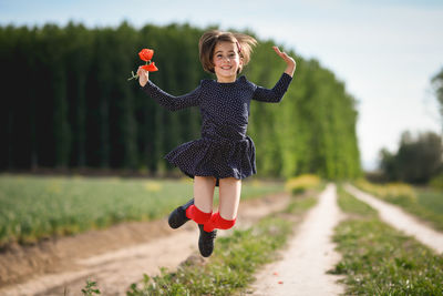 Portrait of girl with flower jumping over field against trees