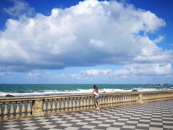 Rear view of man standing on retaining wall by sea against sky