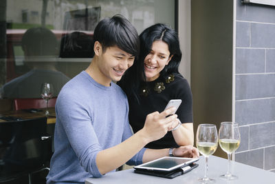 Smiling couple using mobile phone while sitting at sidewalk cafe