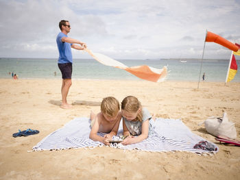 Man holding towel while children sharing smart phone at beach against sky