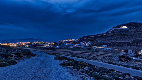 Illuminated road by buildings in city against sky at dusk