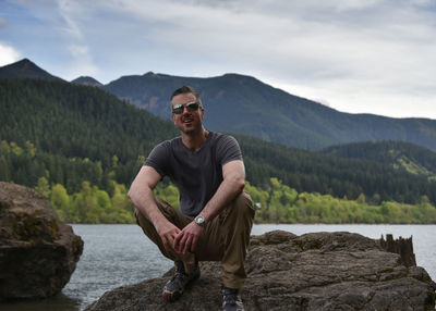 Portrait of man in sunglasses crouching on rock against lake