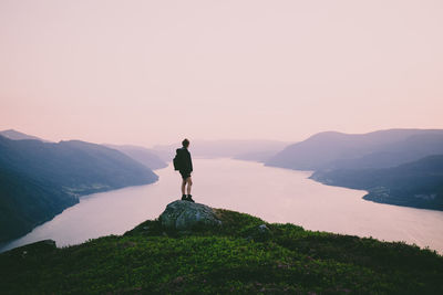 Rear view of woman looking at river amidst mountains against sky during sunset