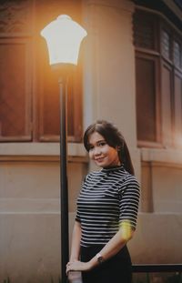 Portrait of a smiling young woman standing against illuminated lamp